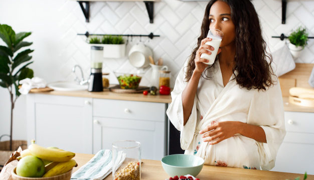 smiled-attractive-mulatto-woman-is-drinking-milk-near-table-with-fresh-fruits-white-modern-kitchen-dressed-nightwear-with-loose-hair-looking-right
